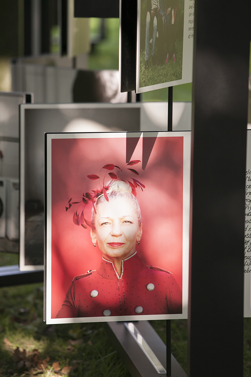 Ein Porträtfoto einer Person in einem roten, verzierten Outfit mit passender Haardekoration. Das Bild ist Teil einer Außenausstellung, die auf einem Metallrahmen montiert ist, und wird von Sonnenlicht angestrahlt. Die Ausstellung befindet sich auf einer Wiese und umfasst weitere Fotos. Kontext: Mögliche Verbindung zu Themen wie Arbeitsschutz und Prävention im Rahmen einer Veranstaltung der Unfallkasse NRW.