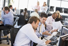 Mehrere Personen arbeiten an Computer-Arbeitsplätzen in einem Großraumbüro. Zwei Männer im Vordergrund betrachten und diskutieren Dokumente, während sie an Computern arbeiten. Andere Mitarbeiter sind ebenfalls an Schreibtischen mit Telefonen und Monitoren beschäftigt. Im Hintergrund stehen weitere Personen und besprechen vermutlich Arbeitsthemen. Die Szene vermittelt einen geschäftigen Büroalltag, der für den Kontext der Unfallkasse NRW relevant sein könnte, insbesondere in Bezug auf Arbeitsschutz und Prävention in Büroumgebungen.