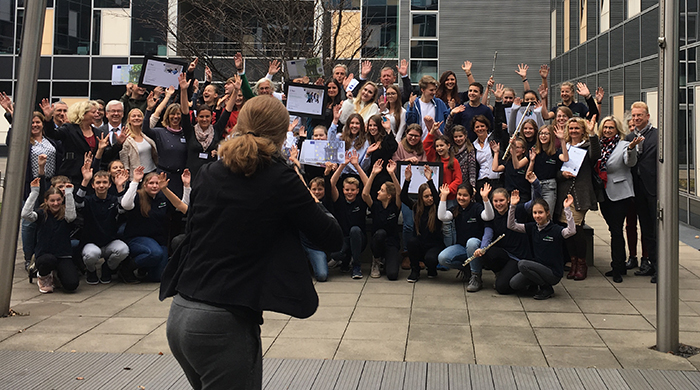 Eine große Gruppe von etwa 50 Personen steht auf einem gepflasterten Hof vor modernen Bürogebäuden. Sie posieren fröhlich für ein Foto, viele halten Zertifikate oder Broschüren in den Händen. Einige Personen tragen Arbeitskleidung oder Uniformen, was auf eine berufsbezogene Veranstaltung hinweist. Im Vordergrund macht eine Person mit dem Rücken zur Kamera ein Foto von der Gruppe. Die Veranstaltung scheint im Zusammenhang mit Arbeitsschutz und Prävention zu stehen, passend zum Kontext der Unfallkasse NRW.