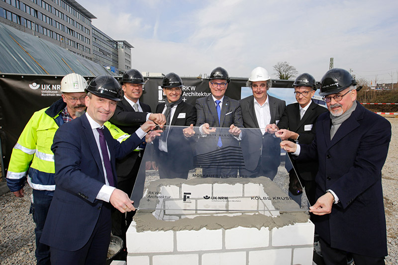 Eine Gruppe von acht Personen mit Sicherheitshelmen beteiligt sich an einer feierlichen Grundsteinlegung. Sie umgeben einen Grundstein aus Mauerwerk und halten gemeinsam eine transparente Platte mit der Inschrift 'ARBEIT. SICHTEN.' Eines der Gebäude im Hintergrund ist mit dem Logo der Unfallkasse NRW gekennzeichnet. Das Foto betont Aspekte des Arbeitsschutzes und der Prävention im Zusammenhang mit der Bauindustrie.