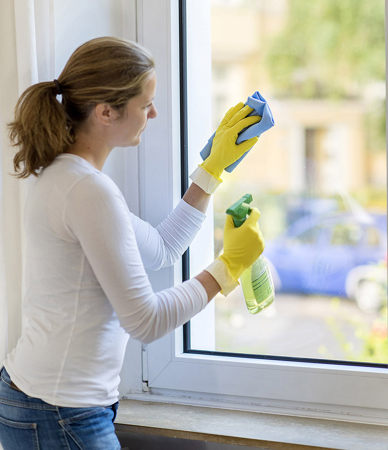 Eine Frau trägt gelbe Gummihandschuhe und reinigt ein Fenster mit einem blauen Tuch und einer Sprühflasche. Sie ist in einem hell beleuchteten Raum, durch das Fenster sieht man verschwommen ein Auto und Grünflächen. Die Szene betont Arbeitsschutzmaßnahmen und Präventionsbewusstsein durch den Gebrauch von Schutzhandschuhen.