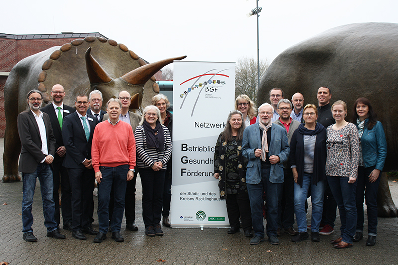 Eine Gruppe von etwa 18 Personen steht vor einer großen Skulptur und einem Banner im Freien. Das Banner trägt den Titel 'Netzwerk Betriebliche Gesundheitsförderung der Städte und des Kreises Recklinghausen' und zeigt das Logo der Unfallkasse NRW, sowie anderer Organisationen. Die Personen sind bunt gekleidet und schauen in die Kamera, was auf ein Gruppenfoto hindeutet. Der Kontext deutet auf eine Aktion zur Gesundheitsförderung und Prävention am Arbeitsplatz hin.