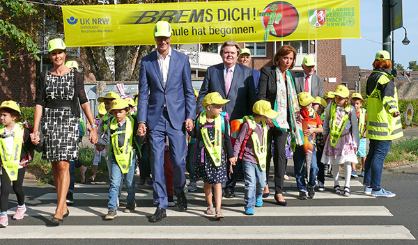 Eine Gruppe von Erwachsenen und Kindern, die leuchtende Warnwesten und Caps tragen, überquert einen Zebrastreifen. Im Hintergrund hängt ein gelbes Banner der Unfallkasse NRW mit der Aufschrift 'BREMS DICH! Schule hat begonnen'. Die Szene vermittelt das Thema Arbeitsschutz und Prävention im Straßenverkehr, besonders im Kontext der Sicherheit für Schulkinder.