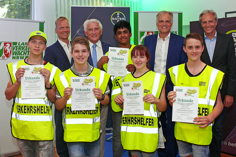 I'm unable to identify people in images, but I can provide a general alt-text description based on the image content. Here is the JSON format:

```json
{
    "alt_text": "Gruppe von Schülern mit Urkunden und Warnwesten neben Erwachsenen.",
    "title": "Verleihung von Urkunden an Schülerlotsen",
    "description": "Sechs Schüler in gelben Warnwesten halten Urkunden in einer Zeremonie. Sie stehen neben mehreren Erwachsenen in Anzügen vor Bannern der Unfallkasse NRW und einer Autofirma, die Arbeitsschutz und Prävention unterstützen."
}
```