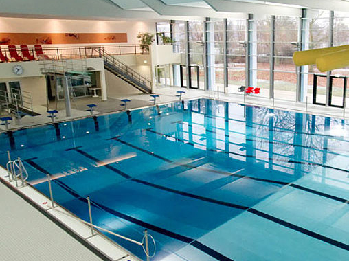 Das Bild zeigt die Innenansicht eines Schwimmbads mit einem großen, leeren Wasserbecken. Im Hintergrund befinden sich große Fenster, die Tageslicht hereinlassen, sowie rote Liegestühle auf einer Galerie. Das Schwimmbad ist sauber und ordentlich, mit Edelstahlleitern und Geländern für sicheren Zugang. An der Seite hängt eine Uhr. Ein gelber Teil einer Rutsche ist im oberen rechten Bereich sichtbar. Das Bild könnte im Kontext des Arbeitsschutzes darauf hinweisen, wie wichtig eine gut gestaltete und gepflegte Einrichtung für die Prävention von Unfällen im Schwimmbadbereich ist.