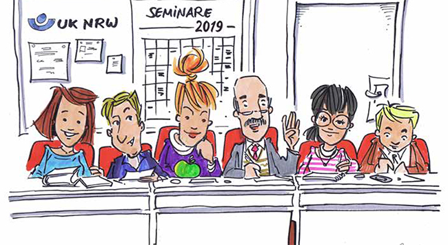 Fünf Personen sitzen lächelnd an einem langen Tisch in einem Seminarraum der Unfallkasse Nordrhein-Westfalen. Hinter ihnen ist ein Kalender mit der Aufschrift 'SEMINARE 2019' an der Wand zu sehen. Die Gruppe ist vielfältig gekleidet und wirkt engagiert. Im Hintergrund befinden sich auch Informationsposter und ein Logo der UK NRW an der Wand. Der Kontext deutet auf eine Besprechung oder Schulung zum Thema Arbeitsschutz und Prävention hin.