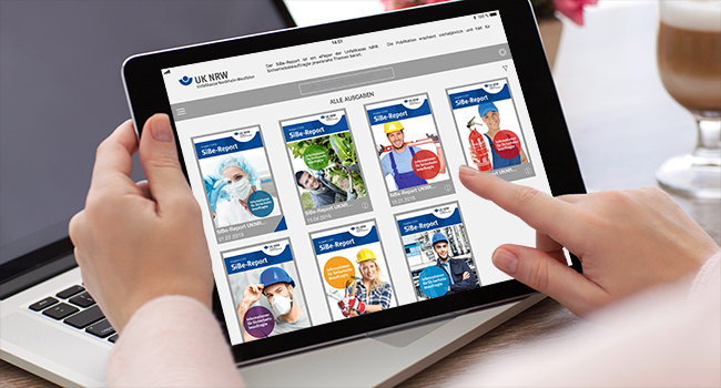 Eine Person hält ein Tablet in der Hand, auf dem Ausgaben des Site-Report Magazins der Unfallkasse NRW angezeigt werden. Jede Magazin-Ausgabe auf dem Bildschirm zeigt ein Bild von Personen in verschiedenen Arbeitsschutz- und Präventionsszenarien, wie z.B. in Sicherheitskleidung. Im Hintergrund ist teilweise ein Laptop und verschwommenes Büroumfeld sichtbar. Der Kontext des Bildes ist die Förderung von Arbeitssicherheit und Prävention durch die Unfallkasse NRW.