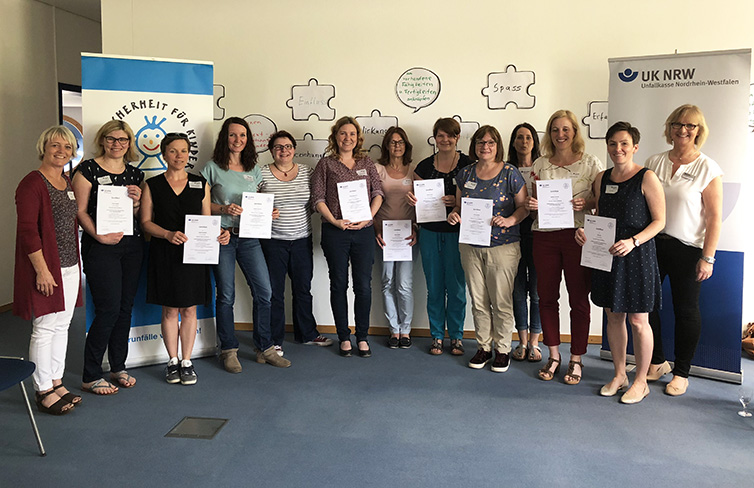 Eine Gruppe von zwölf Personen steht in einem Raum vor zwei Bannern der Unfallkasse Nordrhein-Westfalen. Alle Personen halten Zertifikate in den Händen und lächeln in die Kamera. Im Hintergrund sind weiße Tafeln mit Begriffen wie 'Arbeit', 'Gesundheit' und 'Sinn' an der Wand angebracht. Die Szene suggeriert eine Abschlussfeier oder eine Zertifikatsübergabe im Kontext von Arbeitsschutz und Prävention.