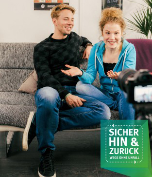 Das Bild zeigt zwei junge Erwachsene, einen Mann und eine Frau, die auf einem Sofa sitzen und lächeln. Sie wirken entspannt und unterhalten sich, während eine Kamera auf sie gerichtet ist. Im Vordergrund befindet sich ein grünes Schild mit der Aufschrift 'Sicher hin & zurück. Wege ohne Unfall', das auf Maßnahmen zur Unfallprävention im Alltag hinweist, ein wichtiges Thema für die Unfallkasse NRW. Das Umfeld deutet auf eine informelle Gesprächssituation hin.