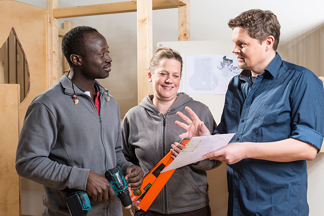 Drei Personen stehen auf einer Baustelle. Zwei von ihnen halten Werkzeuge, während die dritte Person ein Blatt Papier zeigt, das einen Bauplan darstellt. Die Gruppe diskutiert offenbar Sicherheitsmaßnahmen. Im Hintergrund sind Holzwände und Bauzeichnungen sichtbar. Dieses Szenario betont die Wichtigkeit von Kommunikation und Planung zur Gewährleistung der Arbeitssicherheit und Prävention von Unfällen.