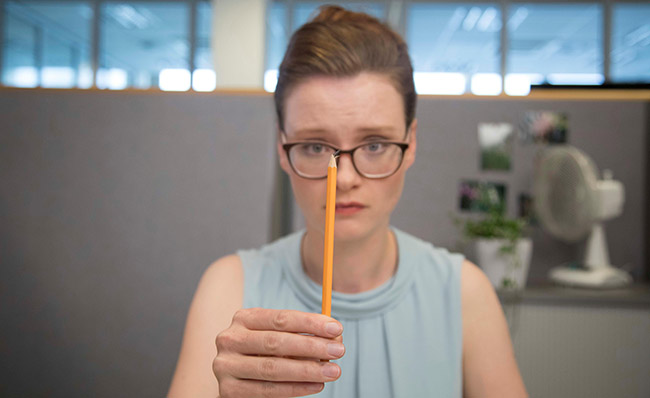 Eine Frau mit Brille sitzt in einem Büro und hält einen Bleistift vertikal vor ihr Gesicht, sodass dessen Spitze auf Höhe ihrer Nase ist. Sie trägt ein ärmelloses, hellblaues Oberteil. Im Hintergrund befindet sich eine graue Trennwand mit leicht erkennbaren Fotos und einem Ventilator, was auf eine Büroumgebung hinweist. Das Bild illustriert das Thema Konzentration und Sehkraft im Kontext von Arbeitsschutz und Prävention bei der Unfallkasse NRW.