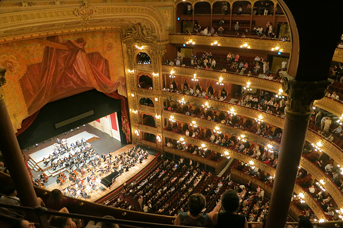 Blick aus der Vogelperspektive auf ein reich verziertes historisches Theater mit roten Samtvorhängen und prunkvollen Balkonen. Im Saal spielt ein großes Orchester auf der Bühne, während die Zuschauer die Aufführung von verschiedenen Etagen aus verfolgen. Die Szenerie ist gut ausgeleuchtet. Der Kontext für die Unfallkasse NRW könnte Arbeitsschutzmaßnahmen bei kulturellen Veranstaltungen betreffen, um Besucher und Künstler zu schützen.