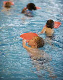 Mehrere Kinder in einem Hallenbad nutzen orangefarbene Schwimmbretter als Hilfsmittel beim Schwimmen. Sie bewegen sich in geordneter Weise durch das Wasser, wodurch Sicherheit und der richtige Umgang mit Schwimmhilfen trainiert werden. Dies fällt unter die Präventionsmaßnahmen der Unfallkasse NRW für sicheres Verhalten im Wasser, besonders im Rahmen von Schulveranstaltungen.