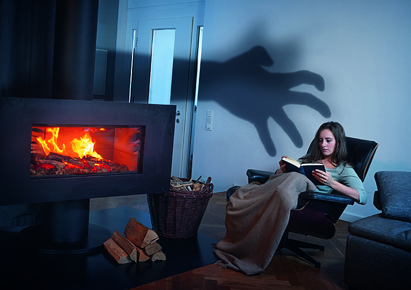Eine Frau sitzt in einem Sessel vor einem modernen, brennenden Kamin und liest ein Buch. Ein Korb voller Feuerholz steht neben dem Kamin. An der Wand hinter ihr ist der Schatten einer großen Hand zu sehen, als ob sie nach ihr greifen würde. Diese Darstellung soll auf Sicherheitsrisiken im Haushalt hinweisen, wie sie von der Unfallkasse NRW thematisiert werden, insbesondere die unbemerkten Gefahren beim Umgang mit Heizquellen.