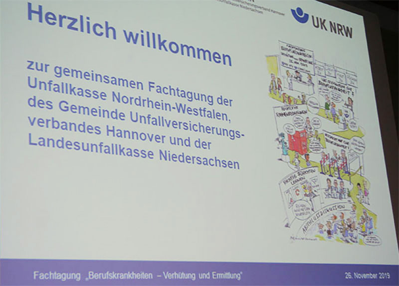 Eine Präsentationsfolie der Unfallkasse NRW mit der Aufschrift 'Herzlich willkommen zur gemeinsamen Fachtagung der Unfallkasse Nordrhein-Westfalen, des Gemeinde Unfallversicherungsverbandes Hannover und der Landesunfallkasse Niedersachsen'. Rechts ist ein Comic abgebildet. Die Veranstaltung trägt den Titel ‚Fachtagung Berufskrankheiten – Verhütung und Ermittlung‘ und findet am 26. November 2019 statt.