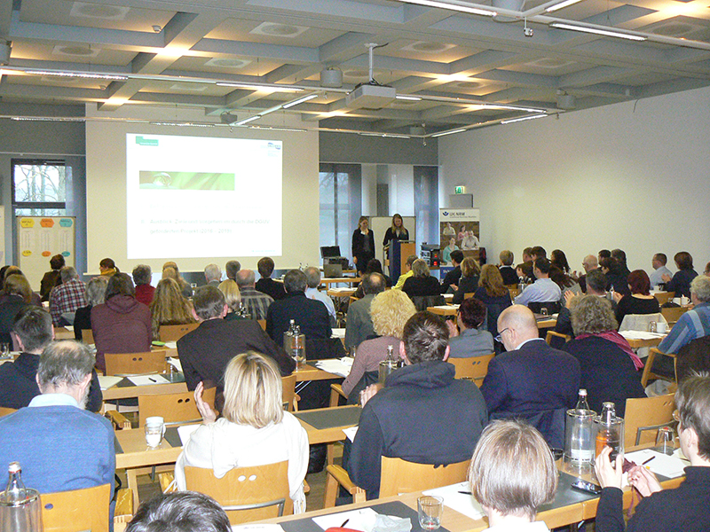 Seminarraum mit mehreren Personen, die einem Vortrag zu Arbeitssicherheit und Prävention bei der Unfallkasse NRW folgen. Zwei Referentinnen stehen vorne neben einer Projektion, die Präsentationsfolien zeigt. Die Zuhörer sitzen an Tischen und hören aufmerksam zu. An den Wänden sind Pinnwände mit Informationen angebracht.