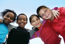 Vier Kinder, zwei Jungen und zwei Mädchen, stehen eng beieinander, lächeln in die Kamera und legen sich freundschaftlich die Arme um die Schultern. Sie symbolisieren Diversität und Gemeinschaft. Der Himmel ist blau im Hintergrund, was eine positive und sichere Umgebung vermittelt. Dieses Bild kann im Kontext von Arbeitsschutz und Prävention für die Förderung von Gemeinschaft und sozialer Unterstützung stehen.