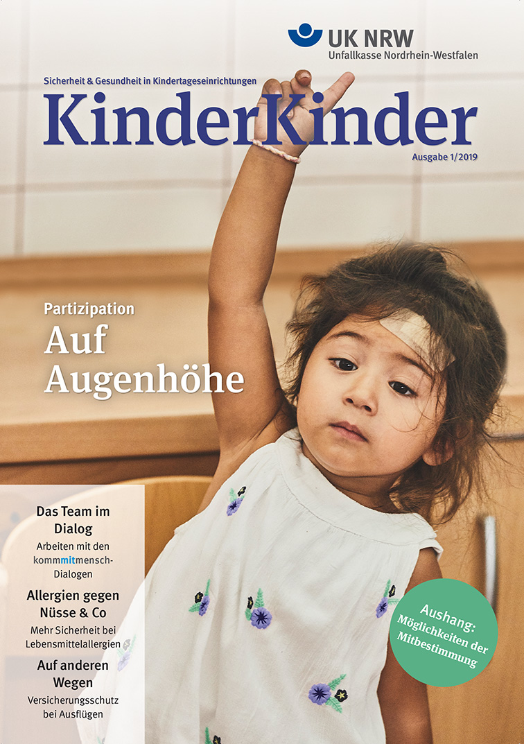 Titelbild der Zeitschrift 'KinderKinder' Ausgabe 1/2019 der Unfallkasse NRW. Ein kleines Mädchen in einer Kindertagesstätte mit einem Pflaster auf der Stirn hebt die Hand, sie trägt ein weißes Kleid mit blauen Punkten. Der Hintergrund zeigt eine Holzwand. Neben dem Mädchen stehen Themen wie 'Partizipation Auf Augenhöhe' und 'Allergien gegen Nüsse & Co'. Der Untertitel der Zeitschrift lautet 'Sicherheit & Gesundheit in Kindertageseinrichtungen'.