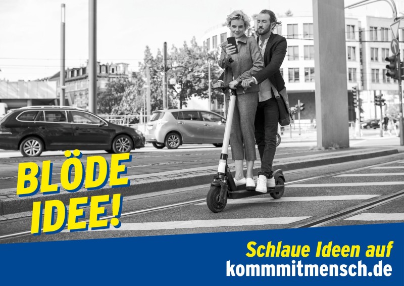 Eine Frau und ein Mann fahren gemeinsam auf einem E-Scooter über eine Stadtstraße. Beide halten ein Smartphone in der Hand und achten nicht auf ihre Umgebung. Der Text 'BLÖDE IDEE!' wird in gelber Schrift über das Bild gelegt. Unten steht 'Schlaue Ideen auf kommmitmensch.de'. Das Bild betont die Gefahren von Ablenkungen im Straßenverkehr und gehört zur Präventionskampagne der Unfallkasse NRW.