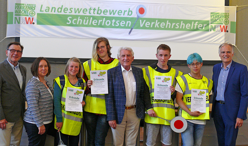 Eine Gruppe von sieben Personen posiert für ein Foto vor einem Banner mit der Aufschrift 'Landeswettbewerb Schülerlotsen Verkehrshelfer NRW'. Vier Jugendliche in gelben Sicherheitswesten halten Zertifikate in den Händen. Zwei Erwachsene stehen auf beiden Seiten der Gruppe. Das Setting deutet auf eine Preisverleihung hin, die die Bedeutung des Arbeitsschutzes und der Prävention bei der Unfallkasse NRW unterstreicht.