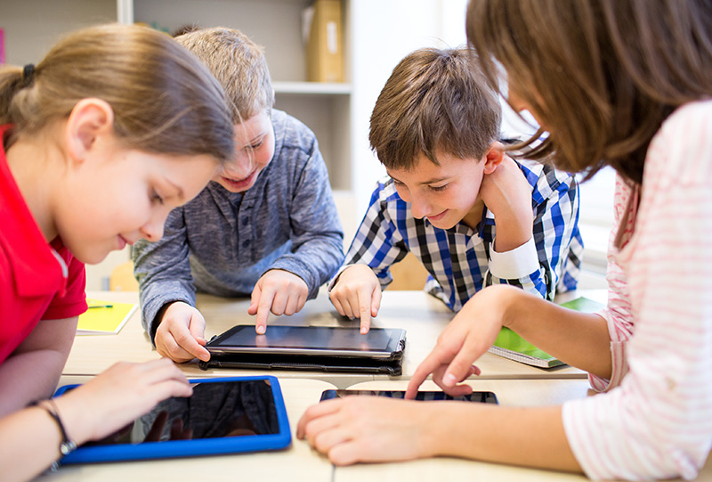 Vier Kinder in einem Klassenzimmer beschäftigen sich konzentriert mit Tablets. Sie sitzen an einem Tisch und scheinen gemeinsam an einer Aufgabe zu arbeiten. Die Szene zeigt die Integration von digitaler Technologie in den Bildungsalltag und betont die Bedeutung von Arbeitsschutz und Prävention durch sicheres und verantwortungsbewusstes Nutzen digitaler Geräte gemäß den Richtlinien der Unfallkasse NRW.
