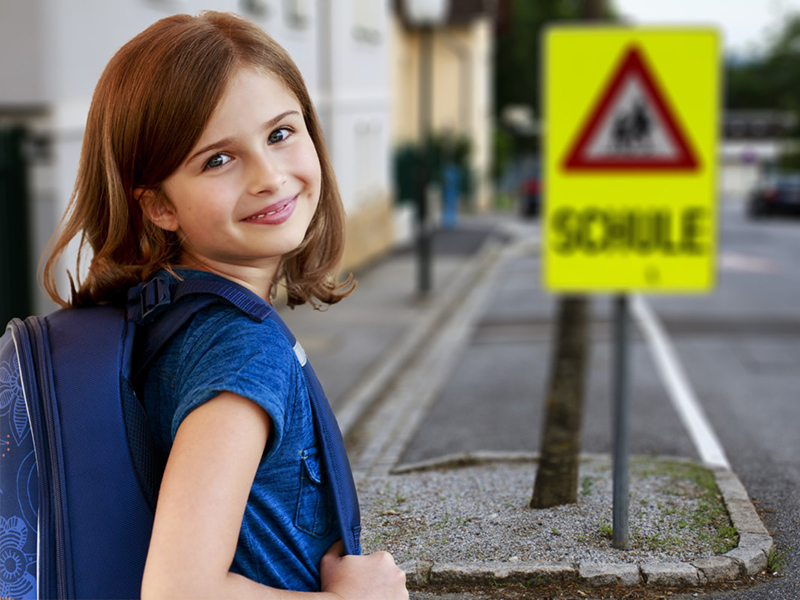 Ein lächelndes Mädchen mit hellbraunen Haaren und blauem Rucksack steht auf einem Gehweg. Im Hintergrund ist ein verschwommenes Schulweg-Schild mit der Aufschrift 'Schule' zu sehen. Diese Szene betont die Wichtigkeit von Verkehrssicherheit und Aufmerksamkeit auf Schulwegen in Bezug auf Präventionsmaßnahmen der Unfallkasse NRW.