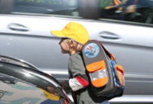 Ein Kind mit einem bunten Schulranzen und gelber Kappe überquert eine Straße vor einem vorbeifahrenden Auto. Das Bild vermittelt die Bedeutung von Schutzmaßnahmen und Sichtbarkeit im Straßenverkehr, um Unfälle zu vermeiden, insbesondere im Zusammenhang mit der Unfallkasse NRW und deren Fokus auf Arbeitsschutz und Prävention.