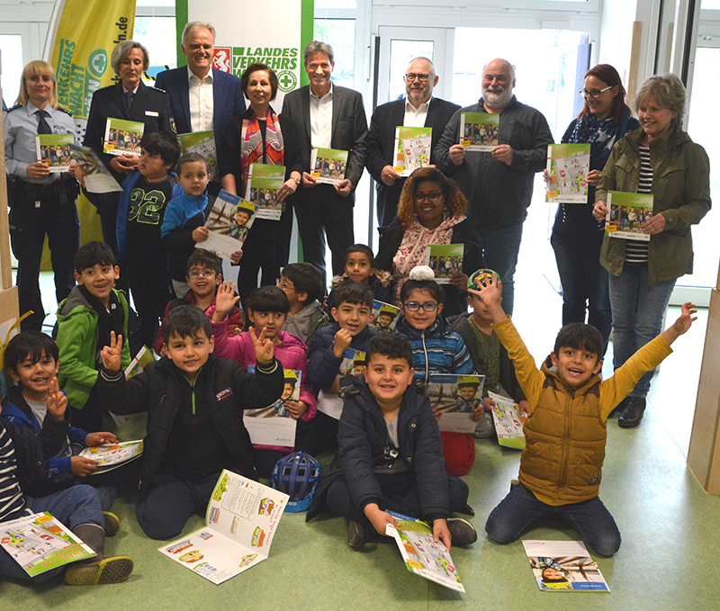 Eine Gruppe von Kindern und Erwachsenen posiert für ein Foto in einem Innenraum. Die Kinder sitzen auf dem Boden und halten bunte Broschüren der Unfallkasse NRW, die sich auf Arbeitsschutz und Prävention konzentrieren. Hinter den Kindern stehen einige Erwachsene mit weiteren Exemplaren der Broschüren. Im Hintergrund befinden sich Banner und Plakate mit Logos der Unfallkasse NRW und anderer Partner. Diese Veranstaltung fördert Themen der Sicherheitserziehung und Prävention bei Kindern im schulischen Bereich.