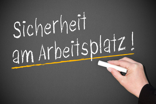 Eine Hand hält ein Stück Kreide und schreibt den Satz 'Sicherheit am Arbeitsplatz!' auf eine dunkelgraue Tafel. Der Satz ist weiß geschrieben und wird durch eine gelbe Linie hervorgehoben. Dies vermittelt die Wichtigkeit von Schutzmaßnahmen und Vorsichtsmaßnahmen am Arbeitsplatz, um Unfälle und Verletzungen zu verhindern. Der Kontext bezieht sich auf Arbeitsschutz und Prävention, besonders wichtig für die Unfallkasse NRW.