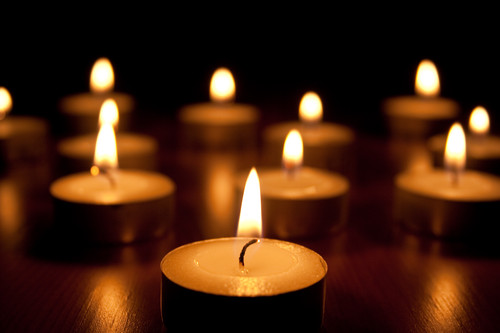 Mehrere kleine Teelichter stehen auf einem dunklen, glatten Untergrund und sind angezündet. Der Raum ist abgedunkelt, wodurch die warme, gelbe Flamme der Kerzen besonders deutlich hervorsticht. Das Bild vermittelt eine ruhige und besinnliche Atmosphäre, die auf mögliche Arbeitsschutzaspekte im Umgang mit offenen Flammen hinweist, wie sie für die Unfallkasse NRW bei Prävention von Bränden relevant sind.