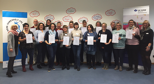 Eine Gruppe von Menschen, bestehend aus 16 Personen, steht in einem Raum und hält stolz Zertifikate in der Hand. Im Hintergrund sind zwei Banner der Unfallkasse NRW zu sehen, die mit ihrem Logo und Slogan das Thema Arbeitsschutz und Prävention betonen. Die Personen lächeln in die Kamera, und an der Wand hängen Blasen mit positiven Schlagworten wie 'Motivation' und 'Teamgeist', die einen unterstützenden und kollegialen Kontext andeuten.