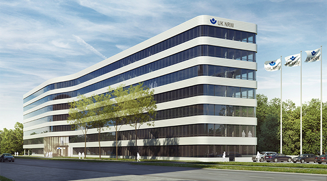 Das Bild zeigt das Hauptgebäude der Unfallkasse NRW mit einer modernen, geschwungenen Architektur. Das Gebäude hat mehrere Stockwerke und ist mit großen Fenstern versehen. Vor dem Gebäude wehen mehrere Fahnen mit dem Logo der Unfallkasse NRW. Im Vordergrund sind Bäume und eine Straße zu sehen. Das Bild vermittelt einen Eindruck von Professionalität und Modernität im Kontext von Arbeitsschutz und Prävention.