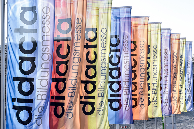 Mehrere an Fahnenmasten befestigte, vertikale Fahnen in verschiedenen Regenbogenfarben. Auf den Fahnen steht 'didacta die Bildungsmesse' in schwarzer Schrift. Der Kontext deutet auf eine Veranstaltung zur Bildung und möglicherweise zur Sicherheits- und Präventionsbildung hin, relevant für Arbeitsschutzthemen der Unfallkasse NRW.