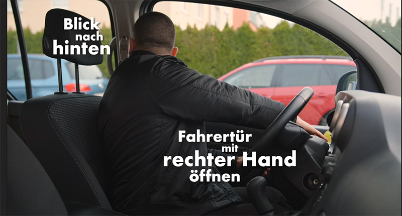 Eine Person im Fahrersitz eines Autos öffnet die Fahrertür mit der rechten Hand, während sie nach hinten schaut. Diese Technik ist Teil von Sicherheitsmaßnahmen, um vorbeifahrende Fahrzeuge oder Fahrräder zu erkennen und Unfälle zu vermeiden. Der Fokus liegt auf der Präventionsarbeit im Rahmen des Arbeitsschutzes. Im Bild sind die Wörter 'Blick nach hinten' und 'Fahrertür mit rechter Hand öffnen' hervorgehoben.