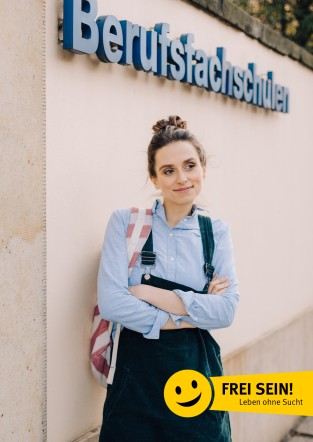 Eine junge Frau mit hochgestecktem Haar steht mit verschränkten Armen vor einem Gebäudewand, an der ein Schild mit der Aufschrift 'Berufsfachschulen' angebracht ist. Sie trägt ein hellblaues Hemd und eine dunkle Latzhose. Auf ihrem Rücken hat sie einen Rucksack. Unten rechts im Bild ist ein gelbes Emblem mit einem lachenden Gesicht und dem Text 'FREI SEIN! Leben ohne Sucht' zu sehen. Das Bild könnte in einem Kontext der Suchtprävention und des Arbeitsschutzes der Unfallkasse NRW stehen.