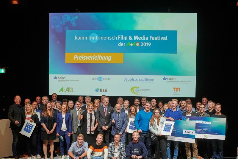 Eine große Gruppe von Menschen steht auf einer Bühne während der Preisverleihung des 'komm mit mensch Film & Media Festival der A+A 2019'. Im Hintergrund ist eine große Leinwand mit dem Logo der Veranstaltung und verschiedenen Logos von Sponsoren und Partnern wie DGUV, Unfallkasse NRW, Basi und anderen zu sehen. Die Teilnehmenden halten Schecks in den Händen, die auf Auszeichnungen und Anerkennungen im Bereich Arbeitsschutz und Prävention hinweisen.