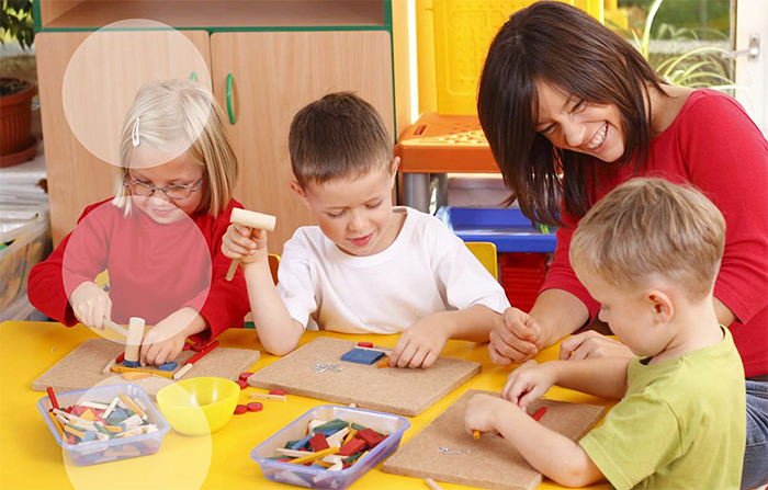 Drei Kinder sitzen an einem gelben Tisch in einem Kindergarten. Sie benutzen kleine Holzhämmer und formen mit bunten Holzspielzeugen. Eine Erzieherin in einem roten Pullover lacht und unterstützt die Kinder. Im Hintergrund sind Regale und Aufbewahrungsboxen zu sehen. Die Szene betont Spiel, Lernen und Betreuung in einer sicheren Umgebung im Kontext des Arbeitsschutzes.