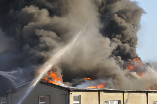Ein großes Feuer brennt auf dem Dach eines flachen Gebäudes. Dichte, schwarze Rauchwolken steigen in den Himmel. Zwei Feuerwehrschläuche spritzen Wasser auf die Flammen. Das Bild vermittelt Dringlichkeit in Bezug auf Brandschutz und Prävention, relevante Themen für die Unfallkasse NRW im Bereich Arbeitsschutz.