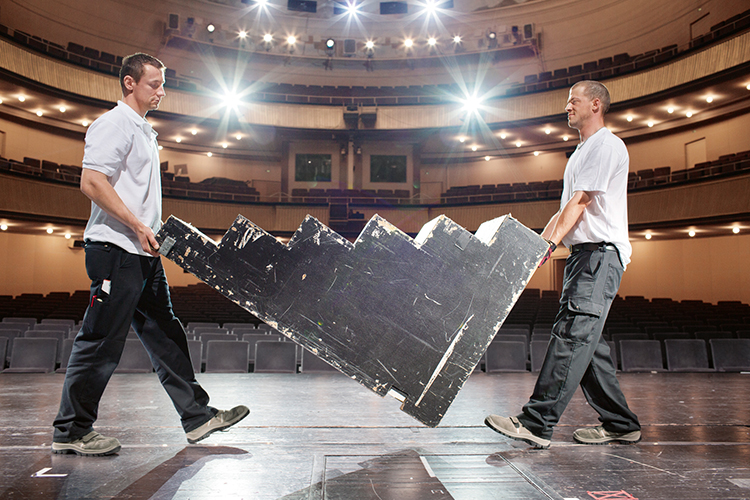 Zwei Männer in Arbeitskleidung transportieren eine große, stufenförmige Platte quer über eine leere Bühne in einem Theater oder Auditorium. Die Bühne ist gut beleuchtet, und sie tragen die Platte gemeinsam an den Enden, um ergonomische Belastung zu minimieren. Die Szene zeigt den sicheren Umgang mit schweren Lasten im Kontext von Arbeitsschutz und Prävention bei Bühnenarbeiten, passend für den Kontext der Unfallkasse NRW.