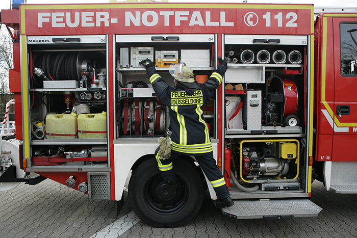 Ein Feuerwehrmann in Schutzkleidung und Helm inspiziert die Ausrüstung an der geöffneten Seite eines Feuerwehrfahrzeugs. Verschiedene Schläuche, Ventile und Geräte sind sichtbar. Auf der Kleidung des Feuerwehrmannes steht 'Feuerwehr Düsseldorf'. Der Fokus liegt auf der Organisation und Verfügbarkeit von Einsatzmitteln für Notfälle. Das Bild vermittelt wichtige Aspekte des Arbeitsschutzes und der Unfallverhütung im Feuerwehrdienst.