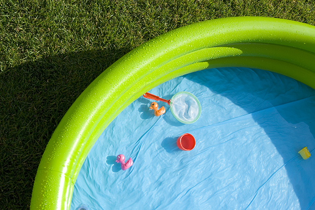 Ein kleiner, aufblasbarer Kinderpool steht auf einer Rasenfläche. Der Pool ist teilweise mit Wasser gefüllt. Darin befinden sich bunte Spielsachen, darunter eine rosa Ente und ein oranger Eimer. Ein kleines grünes Netz zum Schöpfen liegt am Rand. Der Rasen um den Pool ist nass. Der Fokus liegt auf sicherem Umgang mit Wasser im Garten im Kontext von Unfall- und Arbeitsschutz.