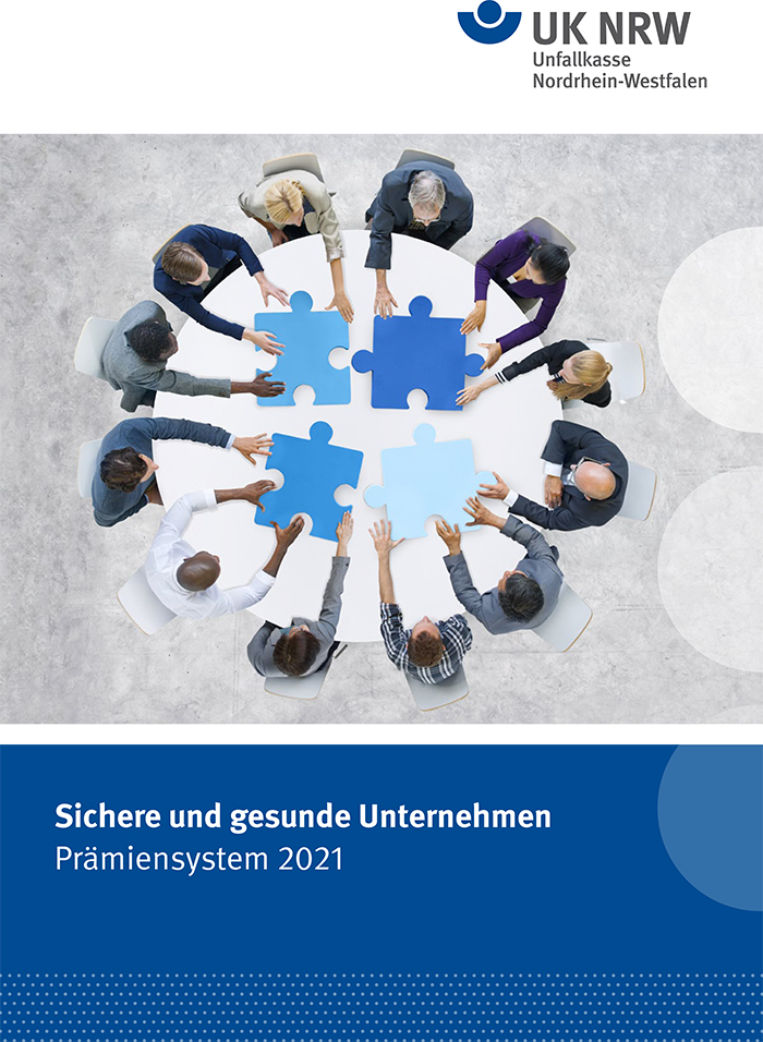 Das Bild zeigt eine Gruppe von zehn Personen unterschiedlicher Demografie, die an einem runden Tisch sitzen und große, blaue Puzzleteile zusammenfügen. Jeder trägt formelle Kleidung. Der Tisch symbolisiert Zusammenarbeit und Prävention im Arbeitsschutz. Im oberen Bereich ist das UK NRW Logo zu sehen. Der untere Bereich des Bildes zeigt den Schriftzug 'Sichere und gesunde Unternehmen, Prämienystem 2021' als Hinweis auf Präventionsmaßnahmen im Rahmen des Prämienprogramms der Unfallkasse Nordrhein-Westfalen.