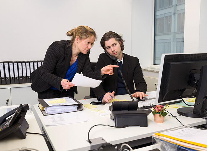 Eine Frau und ein Mann arbeiten in einem modernen Büro. Die Frau zeigt auf ein Dokument auf dem Schreibtisch und erklärt etwas, während der Mann am Telefon ist und den Computerbildschirm betrachtet. Im Hintergrund sind Ordner und ein Fenster zu sehen. Dies unterstreicht die Bedeutung von effektiver Kommunikation und ergonomischem Arbeiten, was für die Prävention von Risiken am Arbeitsplatz wichtig ist, im Kontext der Unfallkasse NRW.