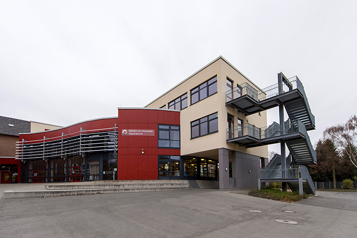 Das Bild zeigt ein modernes Bürogebäude mit einer roten und beige Fassade. An der rechten Seite befinden sich metallene Außentreppen, die Zugang zu den oberen Etagen bieten. Das Gebäude hat große Fenster, die viel Licht hereinlassen. Der graue Himmel deutet auf einen bewölkten Tag hin. Der Gebäudeeingang befindet sich links, und auf der Fassade ist das Logo der Unfallkasse NRW sichtbar. Der Fokus liegt auf sicherer Architektur und Barrierefreiheit.