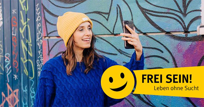 Eine junge Frau trägt einen gelben Hut und einen blauen Pullover. Sie lächelt und macht ein Selfie vor einer bunten Graffiti-Wand. Rechts unten im Bild ist ein gelbes Banner mit einem Smiley und dem Slogan 'Frei sein! Leben ohne Sucht' zu sehen. Diese Darstellung ist Teil einer Kampagne der Unfallkasse NRW zur Prävention von Suchterkrankungen.