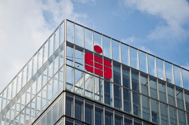 Das Bild zeigt die obere Ecke eines modernen Glasgebäudes mit dem roten, charakteristischen Logo der Sparkasse an der Fassade. Der Himmel im Hintergrund ist teilweise bewölkt. Die klare Struktur des Gebäudes und das markante Logo stehen im Fokus. Im Kontext der Unfallkasse NRW könnte das Bild Themen wie Arbeitssicherheit in Büroumgebungen oder Prävention in modernen Arbeitsplätzen symbolisieren.