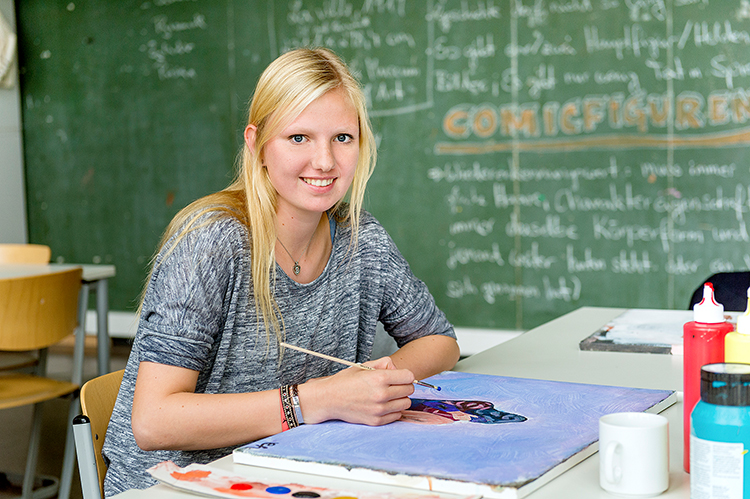 Die Szene zeigt eine junge Frau, die in einem Klassenzimmer an einem Tisch sitzt und ein Bild malt. Sie hat einen Pinsel in der Hand und arbeitet an einem Kunstprojekt. Im Hintergrund ist eine grüne Tafel mit verschiedenen Notizen und dem Wort 'Comicfiguren' deutlich sichtbar. Auf dem Tisch liegen Malutensilien wie Farben und Pinsel. Diese Szene könnte im Kontext der Unfallkasse NRW auf kreative Ansätze zur Prävention und zum Arbeitsschutz in Bildungseinrichtungen hinweisen.