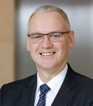 Das Bild zeigt einen lächelnden Mann mittleren Alters in einem dunklen Anzug mit blauer, gestreifter Krawatte und Brille. Er hat kurzes graues Haar und steht vor einem neutralen Hintergrund. Dieses Bild könnte im Kontext von Arbeitsschutz und Prävention bei der Unfallkasse NRW als Beispiel für Professionalität und Vertrauen verwendet werden.