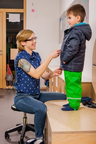 Eine Erzieherin, die auf einem Hocker sitzt, hilft einem kleinen Kind, das auf einem Tisch steht, beim Anziehen seiner Jacke. Das Kind trägt grüne Hosen und Socken. Im Hintergrund ist ein weiterer Raum mit einer Tür sichtbar. Diese Szene zeigt praxisnahes Engagement im Bereich Kinderschutz und Arbeitsschutz in einer Kindertageseinrichtung, relevant für Präventionsmaßnahmen der Unfallkasse NRW.