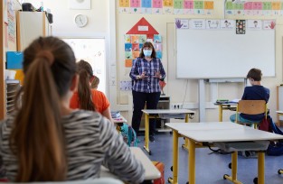 Eine Lehrerin mit Gesichtsmaske steht vor einer Tafel und unterrichtet mehrere Schüler, die auf Abstand sitzen. Die Tische sind paarweise angeordnet. An der Wand hängen bunte Poster und ein Plakat mit geometrischen Formen zeigt Sicherheits- und Verhaltensregeln, was auf Arbeitsschutz und Prävention hinweist, möglicherweise im Kontext der Unfallkasse NRW.